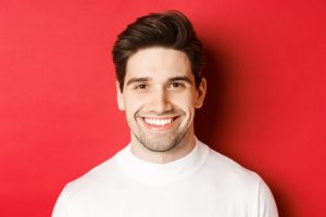 Close-up of handsome smiling brunette man, wearing white sweater, smiling happy and confident, standing against red background